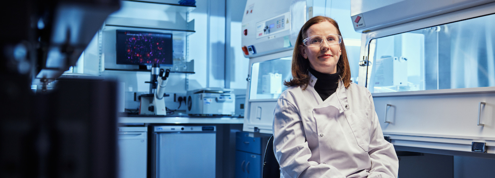 Reseracher sits ina dimly lit lab with clear goggles on, smiling and looking towards the camera